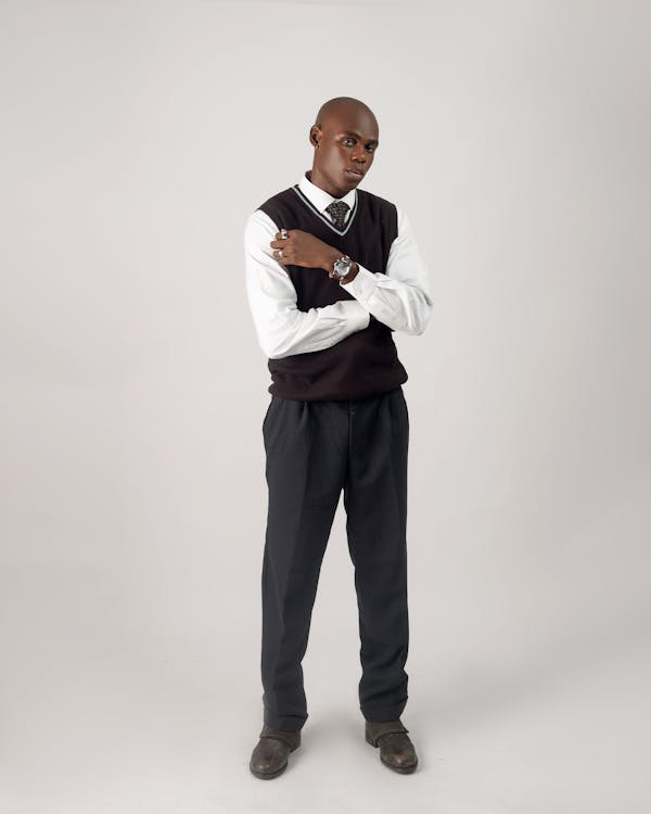 a full body studio portrait of a bald africanamerican man standing with his arms crossed. he is wearing a white dress shirt, a patterned tie, a black sweater vest, black dress pants, and dark colored dress shoes. he also wears a watch and a ring. the background is plain and light.