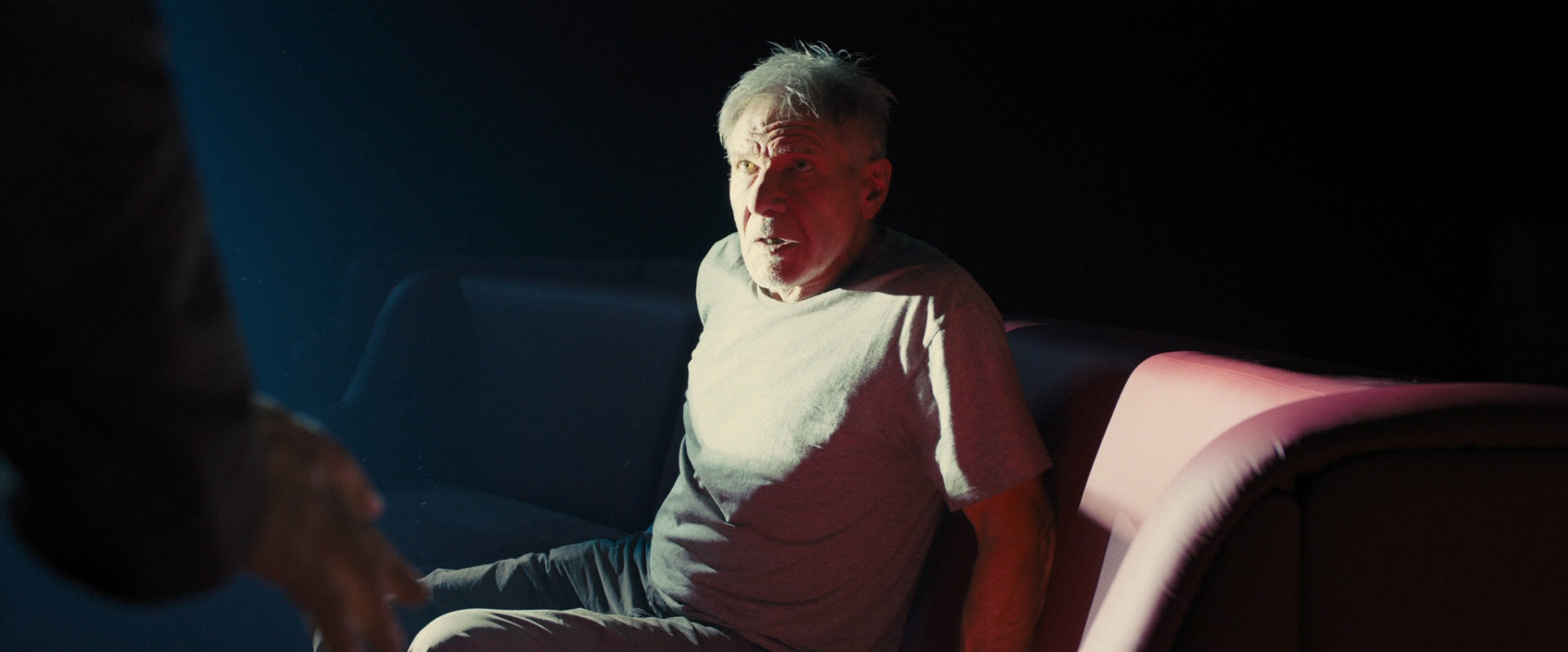 a man sits on a couch in a darkened room, lit by a single light source. he has a worried look on his face and appears to be listening intently to someone offcamera. the man's posture is tense and his expression is serious. the setting suggests a confidential conversation.