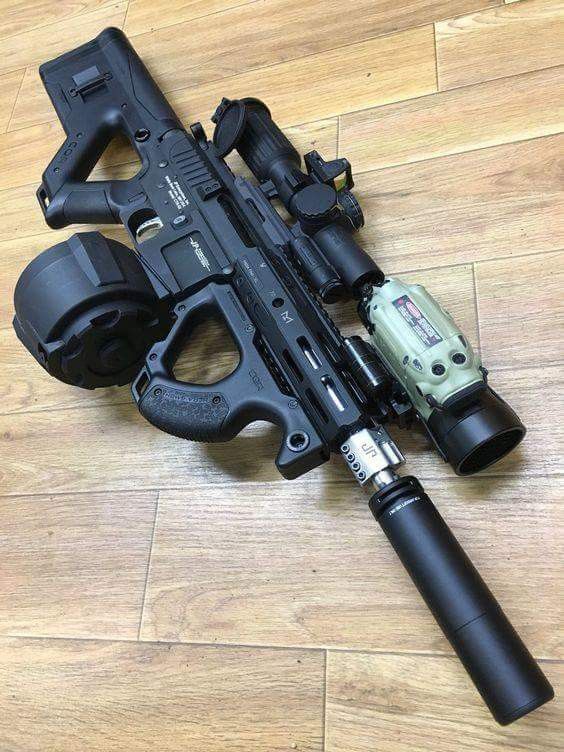a black ar15 pistol is pictured on a wooden floor. it has a drum magazine, an optic, a suppressor, a foregrip, and a weapon light. it also has a pistol brace.