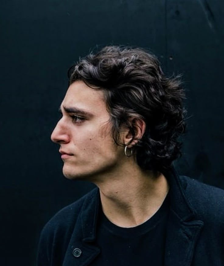 a profile shot of a young man with dark, curly hair. he's wearing a black tshirt and a jacket, and a small hoop earring is visible. the background is a solid dark color.