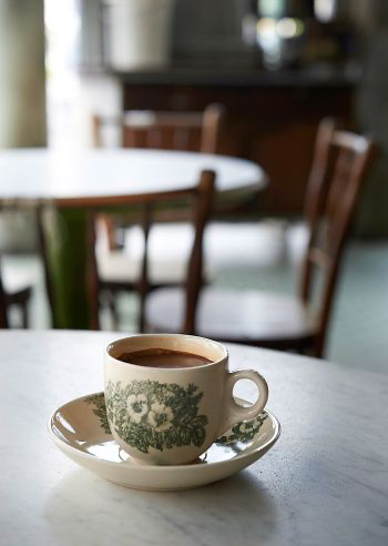 a closeup shot of a white coffee cup with floral pattern and a matching saucer, filled with coffee. the cup is sitting on a table with a blurred background. the background is a cafe with wooden chairs and a table. the cafe's interior is warm and cozy. the coffee looks delicious and inviting.