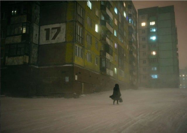a solitary woman in a dark coat walks through a snowcovered street during a blizzard in an urban area with drab apartment buildings. the building on the left has the number 17 painted on its side. many apartment windows glow dimly, providing the only light in the darkness. the scene conveys a sense of cold, isolation, and harsh winter conditions.