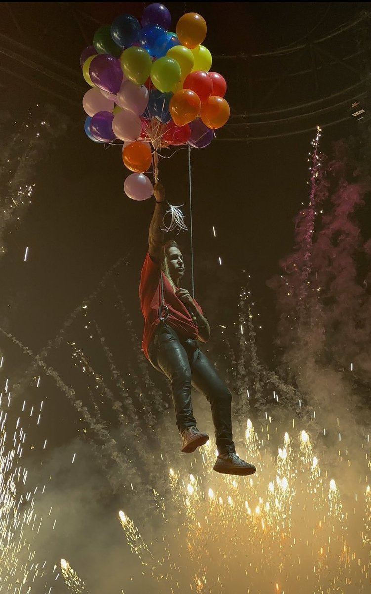 a man in a red shirt and black pants is hanging from a cluster of colorful balloons in the air while fireworks go off in the background. he is likely at a concert or other performance event, and the image captures a moment of joy and excitement. the man's expression is one of happiness and freedom, and the balloons and fireworks seem to symbolize his sense of wonder and optimism.