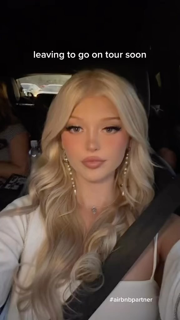 a young woman with blonde hair and makeup is sitting in a car wearing a seatbelt. she is wearing earrings and a necklace. the text on the image says she is leaving to go on tour soon and that she is an airbnb partner.
