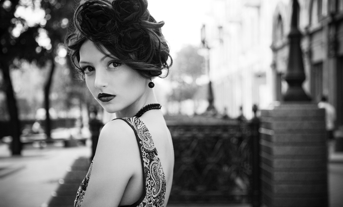 a black and white portrait of a woman in vintage attire. she is wearing a patterned dress, a beaded necklace, and a flower accessory in her updo hairstyle. her dark lipstick emphasizes her lips as she looks over her shoulder towards the camera. the background is blurred, showing an outdoor setting with trees and buildings.