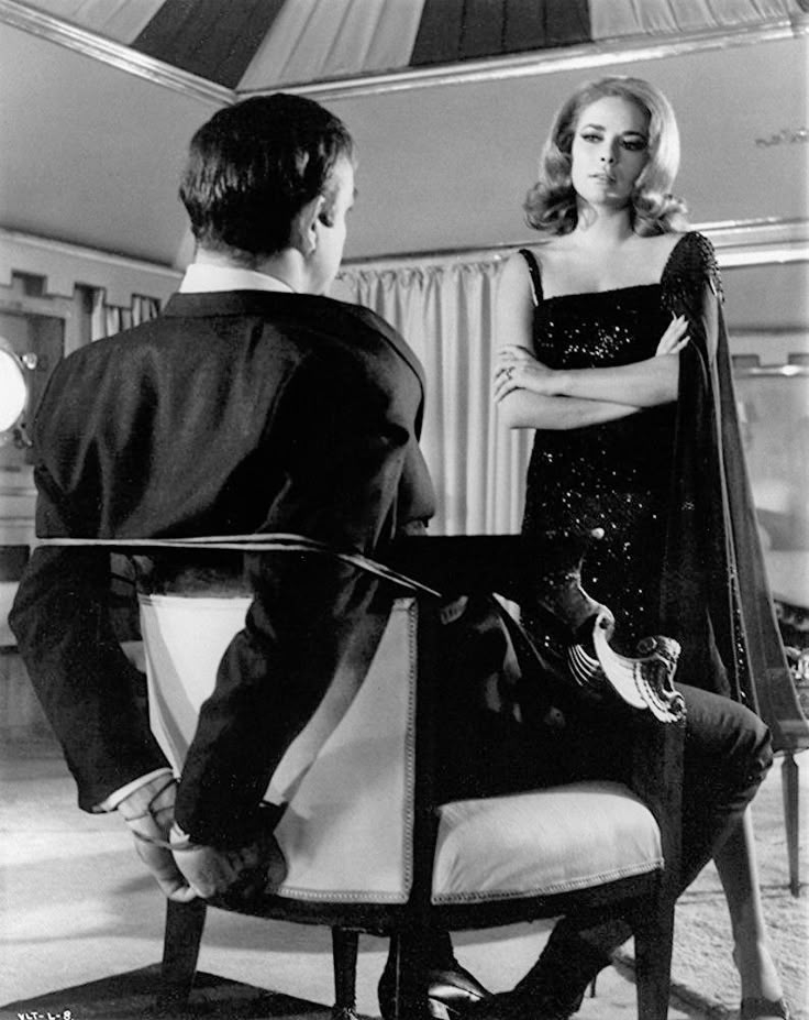 a vintage black and white film still depicts a man tied to a chair with ropes, facing away from the camera. a blonde woman in a sparkly black dress and cape stands with her arms crossed, looking down at him. the scene evokes a sense of suspense and potential danger, typical of a thriller film from the 1960s.