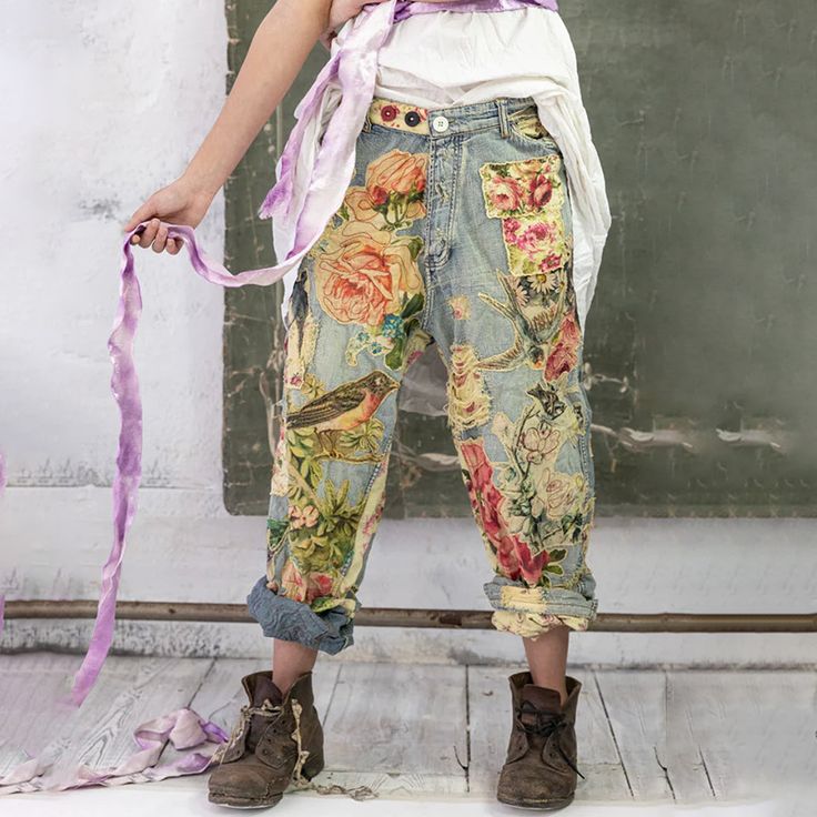a woman stands with her leg slightly bent, showing off her unique fashion style.  she is wearing floral patterned jeans with patchwork and a white shirt tucked into the waistband.  her brown boots, purple ribbon, and white wall add to the unique look.  the model is standing on a wooden floor.
