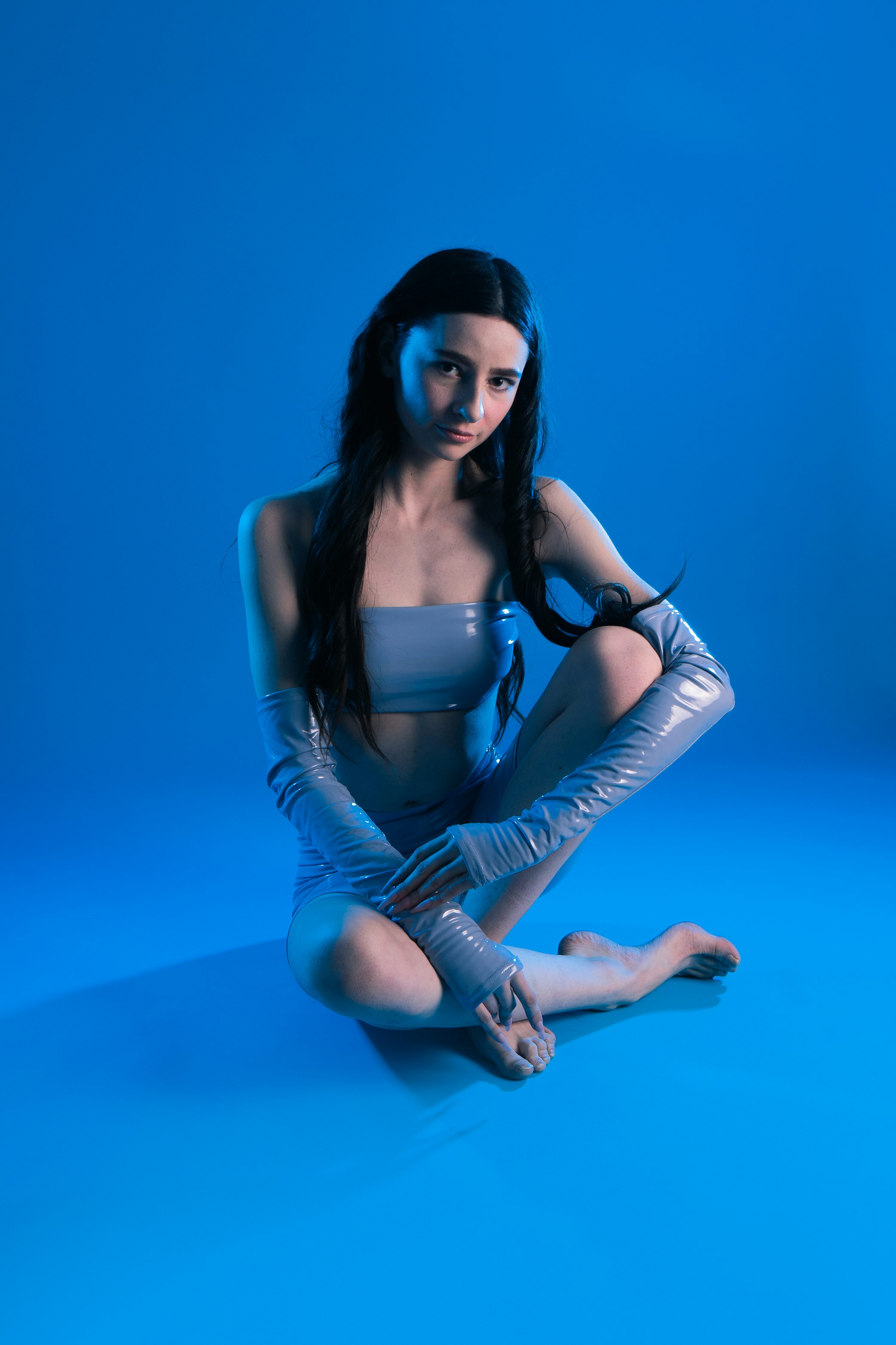 a young woman sits crosslegged on a bright blue background, wearing a light blue bodysuit and matching long gloves. the lighting is predominantly blue, creating a futuristic or cyberpunk aesthetic. she has long dark hair and is looking directly at the camera with a serious expression. the overall style is modern and stylish.