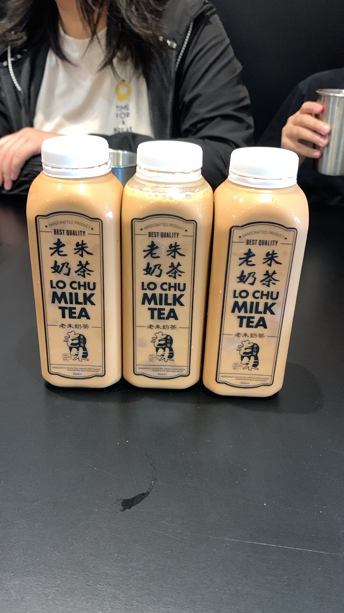 three plastic bottles of lo chu milk tea stand on a dark tabletop. a person wearing a black jacket is partially visible in the background, and another person's hand holding a metal cup is also visible. the bottles are labeled with chinese characters and english text, indicating the brand and flavor of the beverage. the scene appears to be set in an indoor cafe or restaurant.