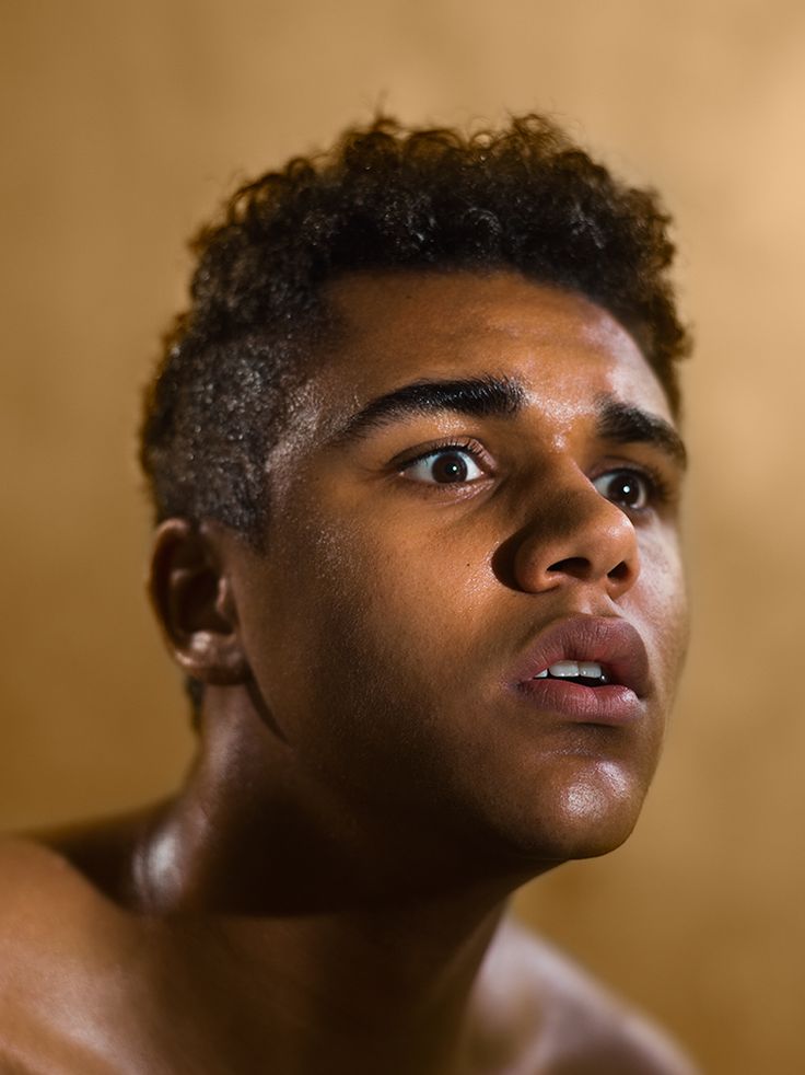 a closeup portrait of a young man with curly dark hair, looking up in shock or surprise with his mouth open and eyes wide. his face is lit from above, casting shadows around his features. his skin is dark and his shoulders are bare. the background is a warm golden yellow.