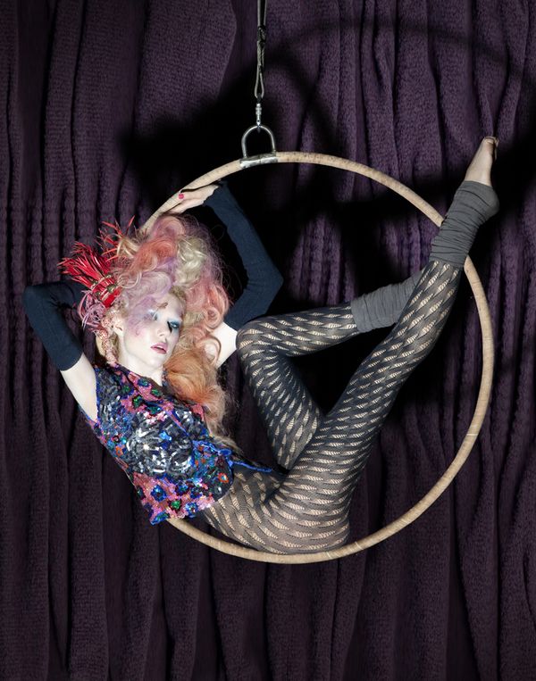 a woman is hanging upside down in a hoop. she is wearing black patterned tights, a sequined blue and pink top, and has pink and blonde hair with red feathers. she is in a studio with a dark purple background.