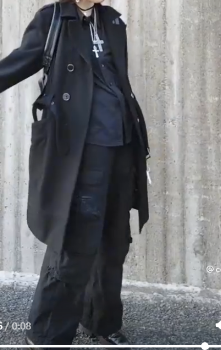 a woman is standing on a street wearing a black trench coat, black cargo pants, and a silver chain necklace. her look is stylish and edgy, with a hint of urban flair.