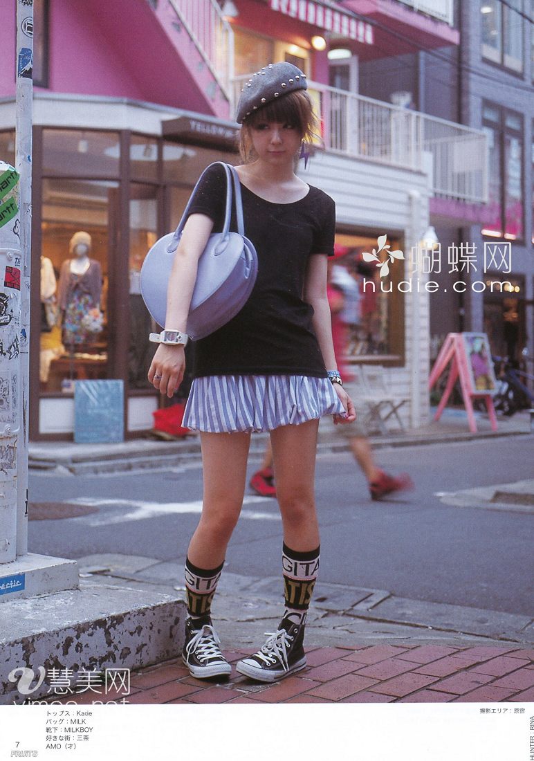 a young woman with short brown hair and a black beret stands on a city sidewalk. she's wearing a black tshirt, blue and white striped skirt, black and white striped socks, and black and white converse sneakers. she's carrying a large, round, light blue purse over her right shoulder. she's posing for a street style photo, with her left hand resting on her thigh and her right hand slightly raised. the background is a busy street scene with shops, buildings, and other people.
