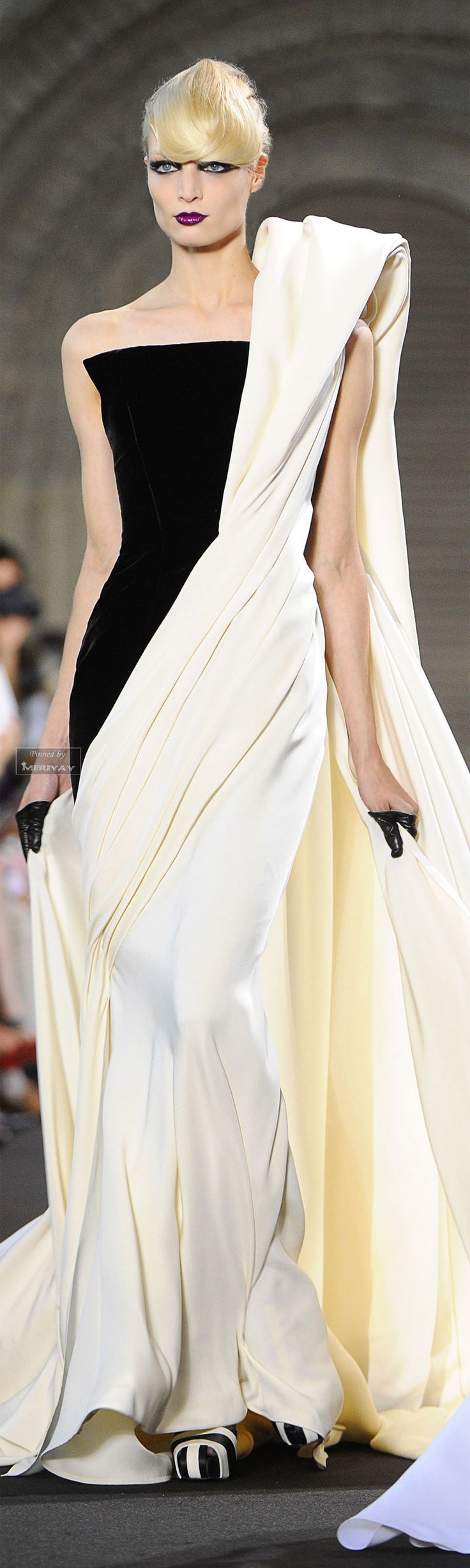 a fashion model wearing a white and black gown stands on a runway looking into the camera. the model is wearing a black velvet bodice and a white satin skirt with a dramatic drape that flows behind her. she is also wearing dark eyeshadow and lipstick, and has her hair styled in a side part with loose waves.  the model's elegant pose and the gown's design highlight the beauty of the clothing. the photo appears to be taken at a fashion show or photoshoot.