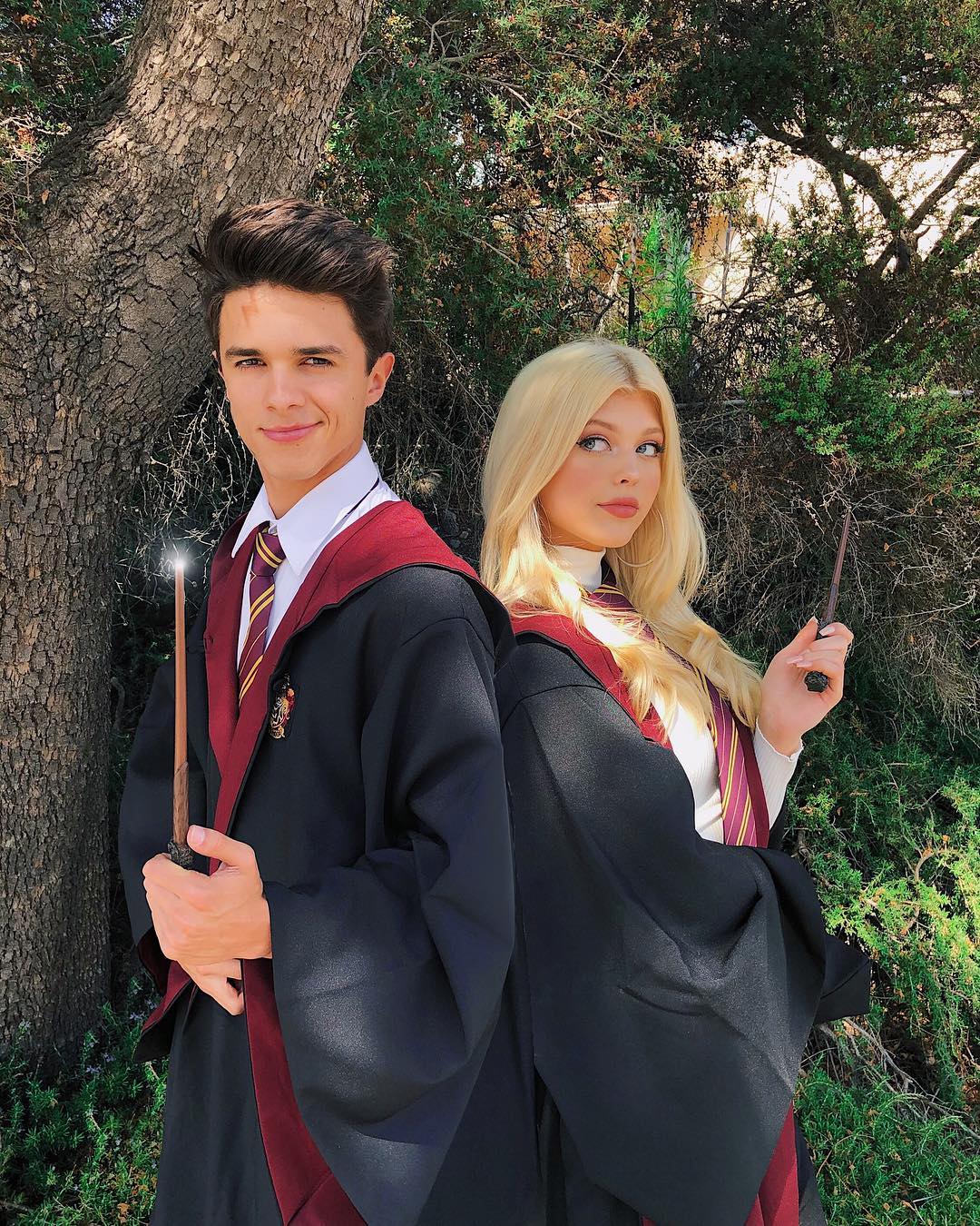 a man and a woman dressed as harry potter characters are standing back to back in front of a tree. the man is on the left, wearing a gryffindor robe, a white shirt, and a gryffindor tie. he is holding a wand in his right hand, which has a faint glow at the tip. the woman is on the right, wearing a similar gryffindor robe, a white turtleneck, and a gryffindor tie. she is holding a wand in her right hand. they are surrounded by greenery, with a tree trunk visible behind the man and various bushes and shrubs in the background. the light is bright, suggesting a sunny day. the man is brent rivera and the woman is loren gray, both are cosplaying as harry potter characters.