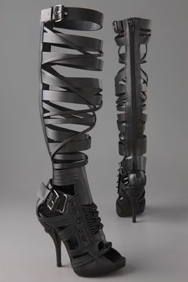 two black highheeled boots are standing next to each other on a gray background. the boot on the left is kneehigh and has a cagelike design made of straps and buckles. the boot on the right is also kneehigh and has a similar cagelike design with a zipper in the back.