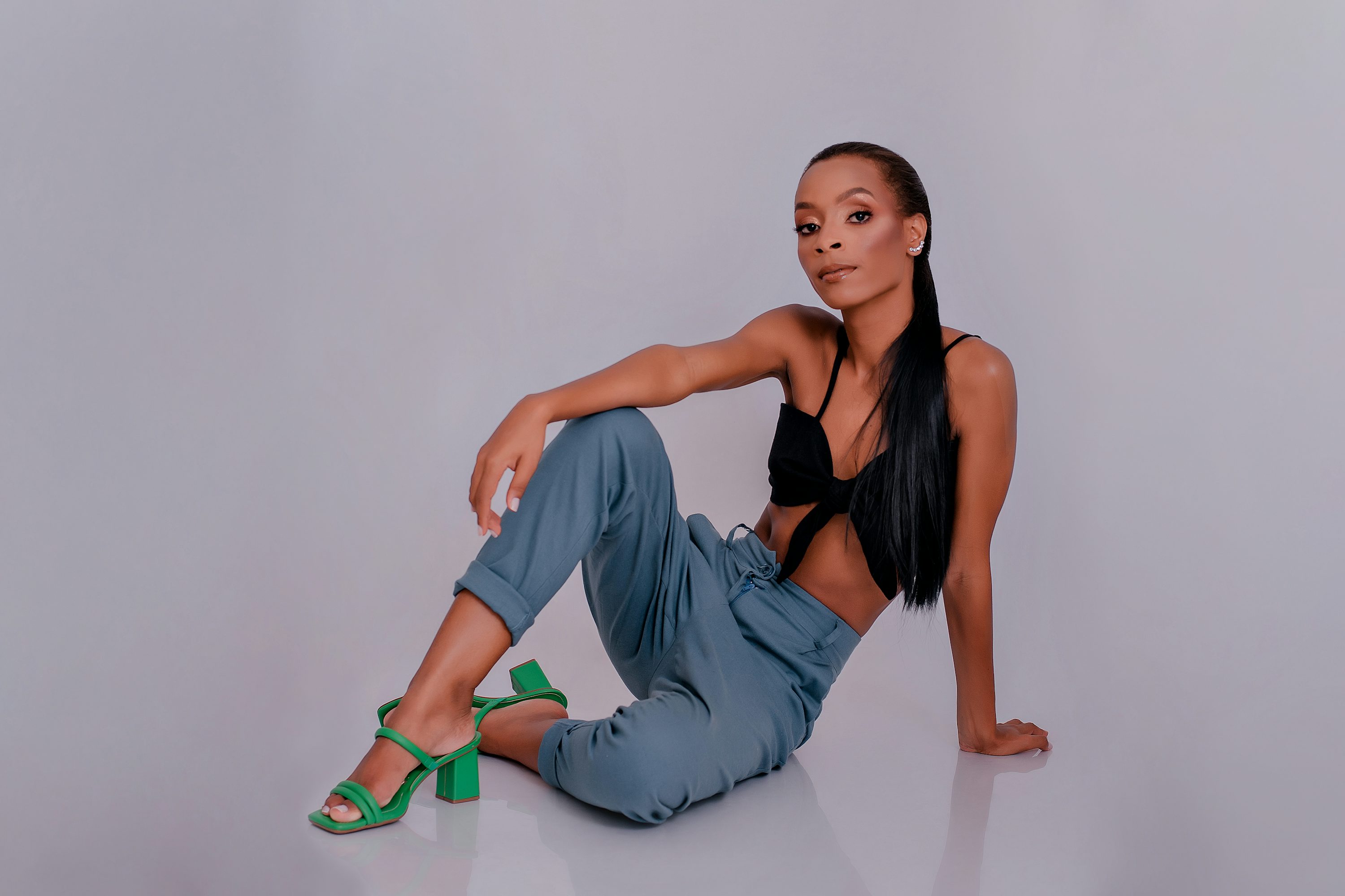 a young, attractive black woman with long black hair is sitting on a grey background. she is wearing a black crop top and loose blue pants, and stylish green highheeled sandals. she is posed confidently, looking at the camera with makeup on.  the overall image is a fashion shot showcasing the model and her outfit.