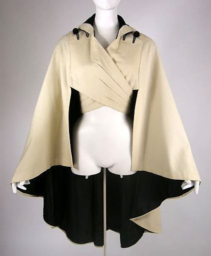 a creamcolored cape coat with a black lining is displayed on a mannequin. the cape has a wide, flowing silhouette and fastens at the neck with a black collar and decorative buttons. the garment is open at the front, revealing the black lining and suggesting it is designed to be worn over a dress or other outfit. the image is likely from a fashion archive or museum collection, highlighting a specific style or era of clothing.