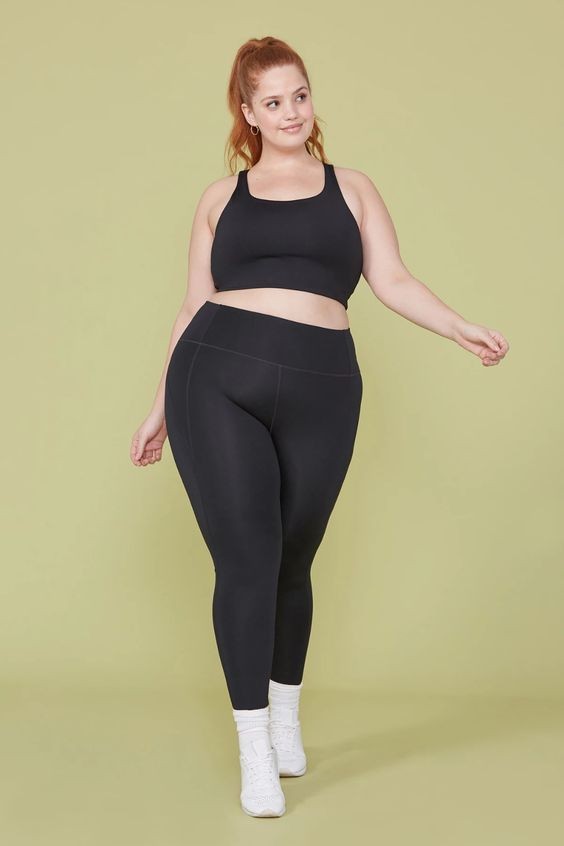 a curvy woman with reddish hair tied back stands in front of a pale green background.  she is modeling a black sports bra and matching black leggings.  she is wearing white ankle socks and white platform sneakers.  she is posing with her left leg slightly forward and her left arm outstretched to the side. her pose and expression are confident and friendly. the lighting is soft and diffused, creating a flattering and approachable feel.