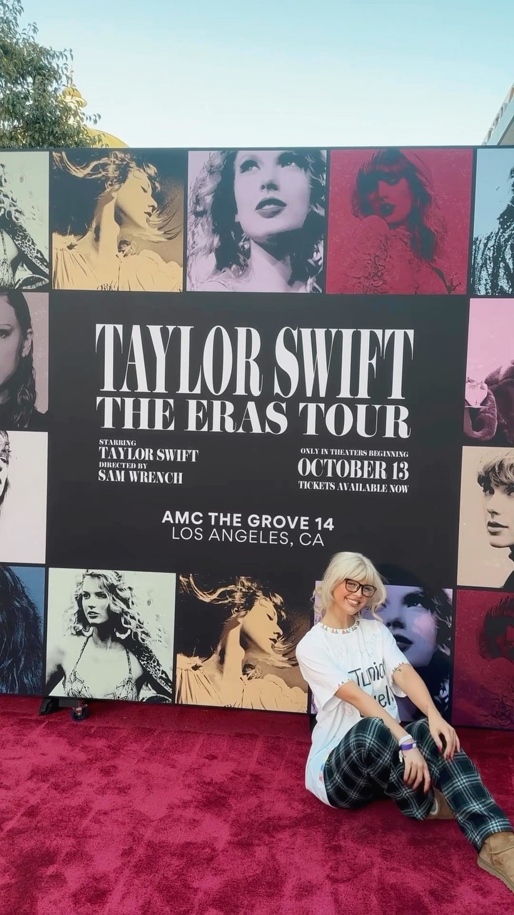 a woman with blonde hair and glasses poses in front of a taylor swift "the eras tour" movie poster. she is wearing a white tshirt and plaid pants. the poster advertises the movie's release date as october 13 at amc the grove in los angeles.