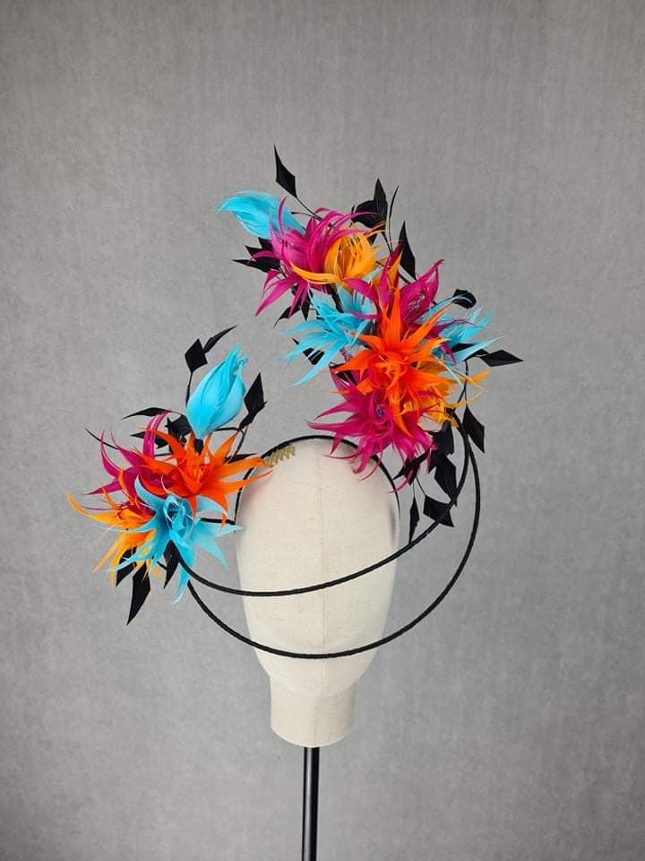 the image shows a colorful and ornate headpiece or fascinator displayed on a mannequin head. the headpiece features vibrant arrangements of feathers in colors such as pink, orange, and turquoise, accented with black decorative elements that resemble leaves or abstract shapes. the headpiece is mounted on a black wire frame that curves around the mannequin's head. the overall impression is dramatic and stylish, suggesting a fashion accessory for special occasions or costume wear. the mannequin is white and displayed on a black stand, set against a gray background.