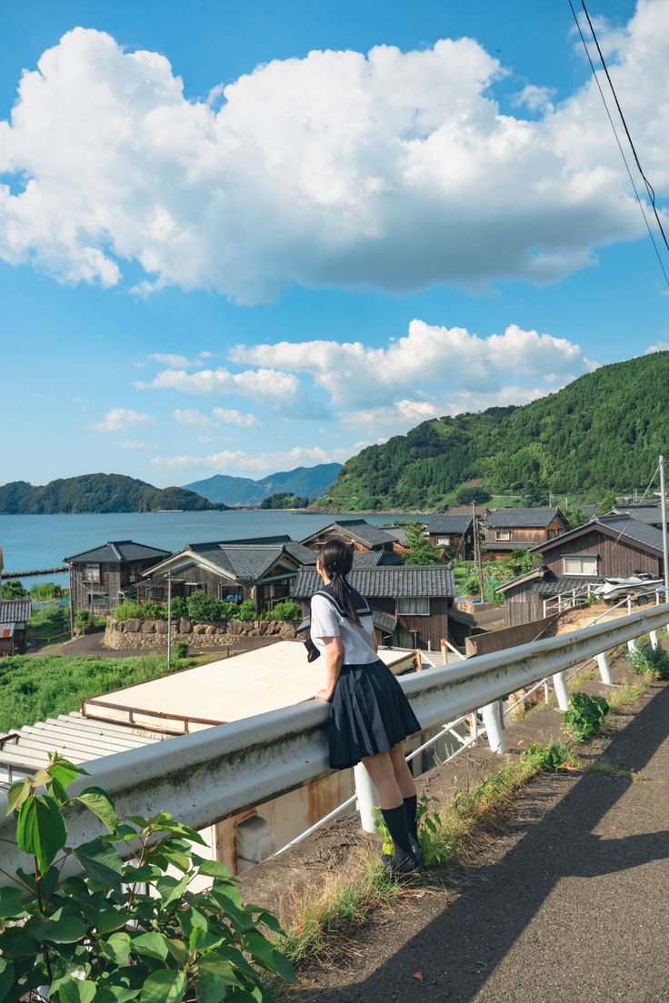 a young japanese schoolgirl in a traditional sailor uniform stands on a guardrail overlooking a picturesque seaside town nestled among mountains. the ocean is visible in the background, and the sky is a clear blue with fluffy white clouds. the scene is peaceful and idyllic, evoking a sense of tranquility and showcasing the beauty of a coastal japanese village. the girl gazes out towards the sea, taking in the beautiful surroundings. the image's color palette is vibrant and harmonious, emphasizing the serene atmosphere.