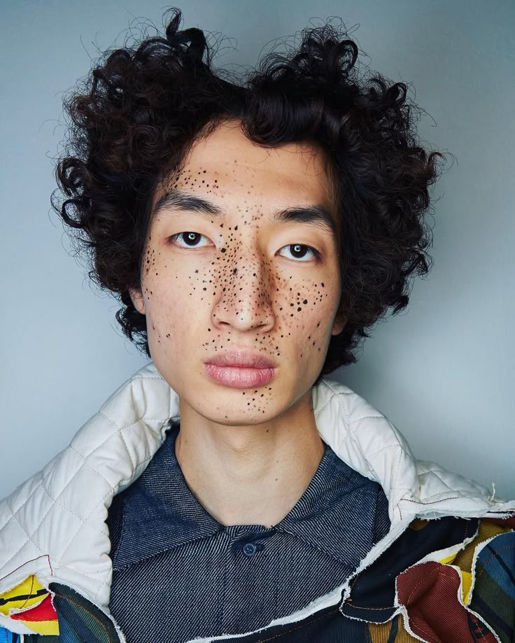 a closeup portrait of an asian man with dark curly hair and numerous freckles on his face. he is wearing a denim shirt and a quilted jacket with a colorful patchwork design. the image is a studio shot with a simple background.