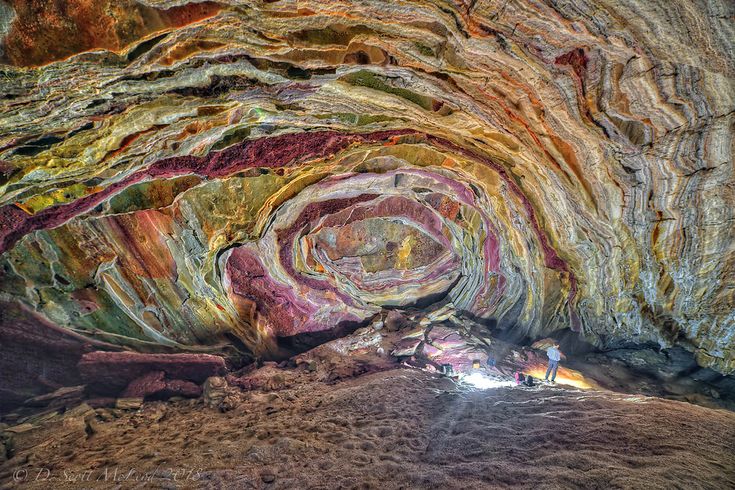 the image shows the interior of a colorful cave. the walls and ceiling are composed of layers of rock with varying colors, including shades of red, yellow, green, and brown. a person stands in the foreground, providing scale to the vastness of the cave. light streams in, illuminating the rocky floor and creating shadows. the rock formations exhibit unique textures and patterns.