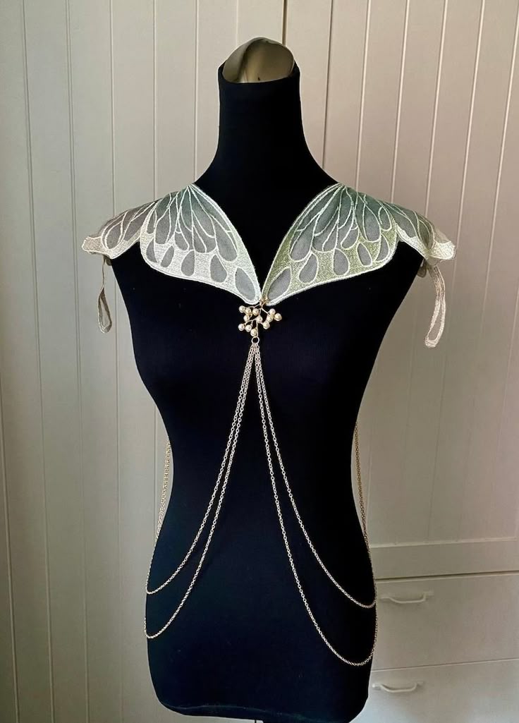 a black mannequin wears a fantasyinspired butterfly collar with a body chain. the collar is designed like butterfly wings, featuring white and green details. a pearl embellishment connects the collar to the draping silver chains that form the body chain. the background consists of a white paneled wall and a white cabinet.