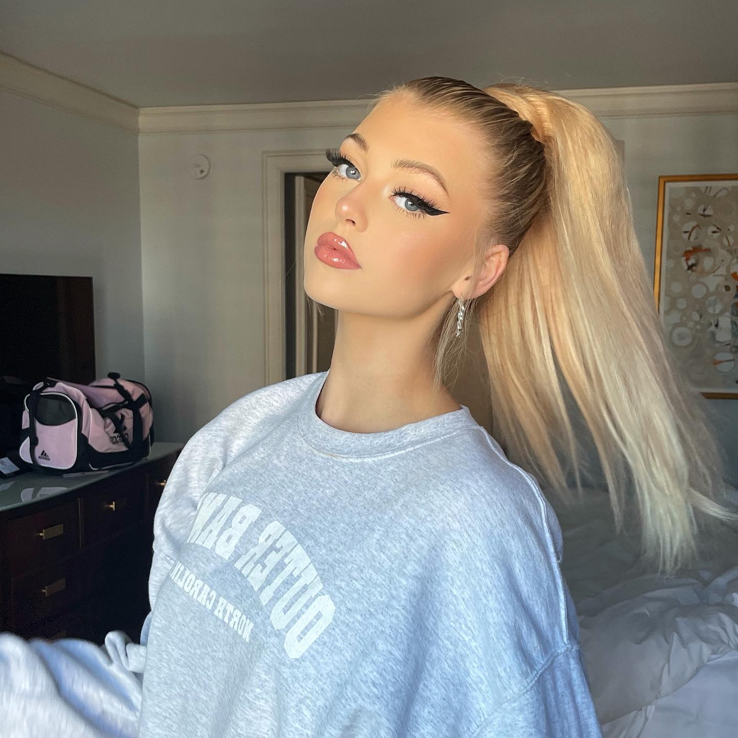 a woman with blonde hair in a high ponytail wearing a gray sweatshirt is posing for a photo in what appears to be a hotel bedroom. she has dramatic eyeliner and earrings. the sweatshirt has "duke by north carolina" written on it in large letters. a luggage bag is on a dresser behind her, and a bed is in the foreground.