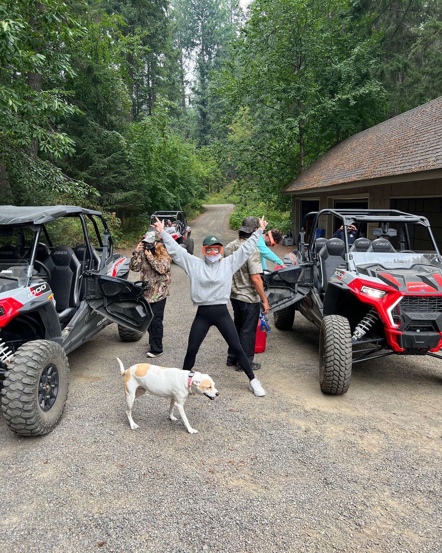 the image shows a group of people gathered near two parked offroad vehicles (likely polaris rzrs), in a wooded area. a woman in a grey sweatshirt is standing with her arms raised in a celebratory pose.  she's wearing a face mask. other people are standing nearby, some appearing to be preparing or getting out of the atvs. a small, white and tan dog stands calmly in the foreground. the background reveals a forested landscape, with a rustic cabinlike structure visible. the overall mood is one of leisure and outdoor adventure.
