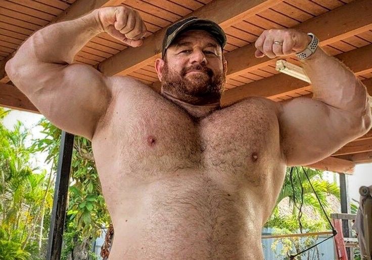 a muscular, hairychested man flexes his biceps and triceps outdoors. he is wearing a baseball cap and has a red beard. the setting appears to be a covered patio or porch area with wooden beams.  the man's physique is very welldefined and he appears to be in excellent physical condition. the image focuses on his impressive musculature.