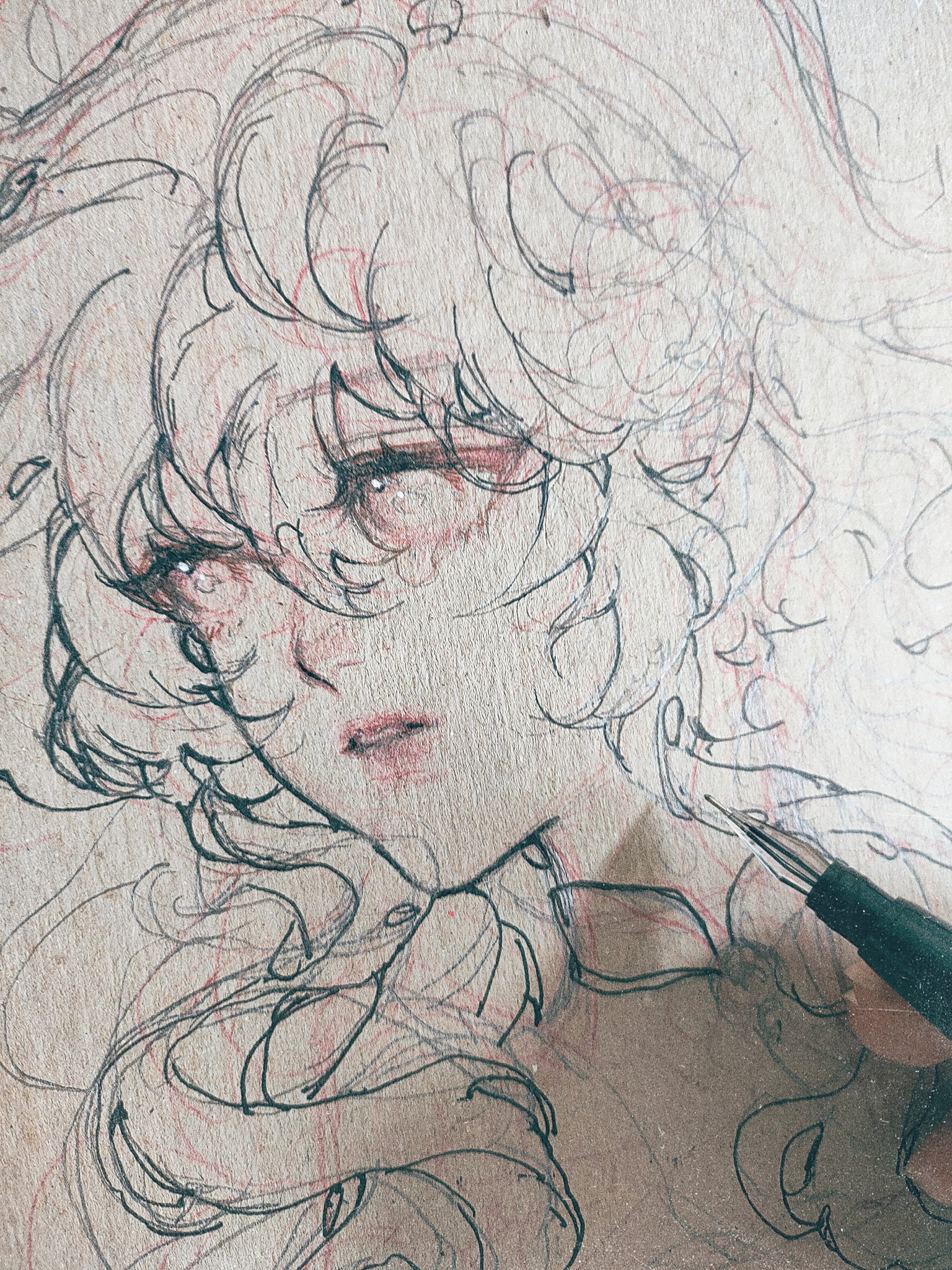 the image is a closeup shot of an artist's hand sketching a detailed animestyle portrait of a girl's face. the sketch is unfinished, with visible pencil lines and shading under the darker ink lines. the girl's hair is voluminous and stylized, and her expression is soft and somewhat melancholic. the background is filled with additional, lighter pencil markings that might be preliminary sketches or guide lines. the artist is using a pen or a finetipped drawing instrument to create the dark lines in the artwork. the paper has a visible texture. the overall style is delicate and emotive.