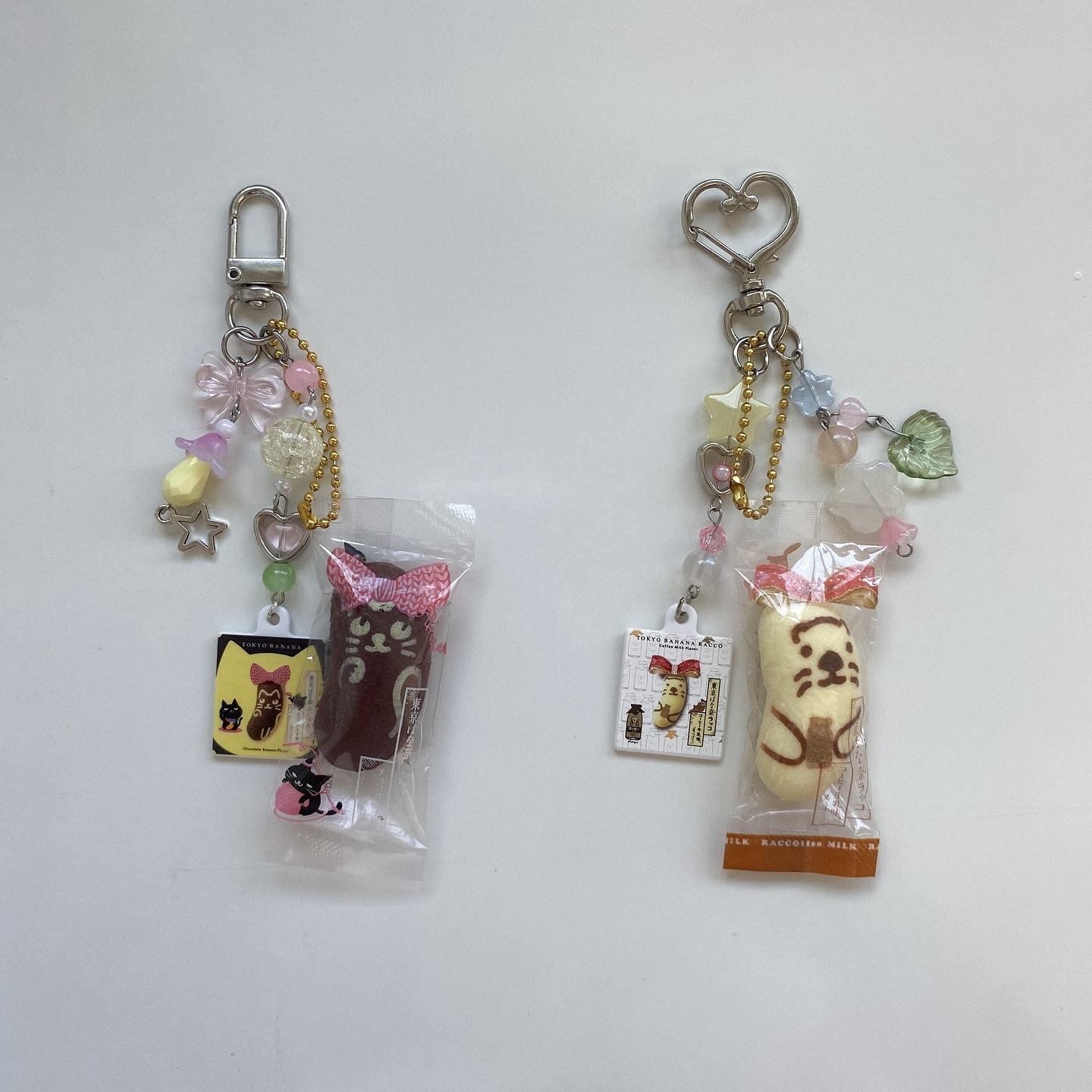 two keychains featuring miniature sweets and decorative charms are displayed against a plain white background. the keychain on the left has a silver clasp, a gold chain, and various pastelcolored beads and charms, including a star and a bow. it's attached to a small, wrapped chocolate snack with a cat design. a rectangular tag with a cat illustration is also part of the keychain. the keychain on the right has a heartshaped clasp and similar pastelcolored charms, along with a gold chain. it's attached to a small, wrapped tokyo banana snack with a sea otter design. the keychain also includes a rectangular tag with a tokyo banana illustration. both keychains have a cute, kawaii aesthetic.
