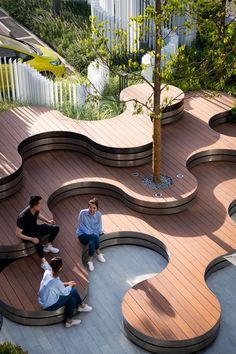 this aerial shot captures a contemporary outdoor public space featuring a wooden deck with a unique curvilinear design. people are seen sitting and interacting on the deck, which is integrated with trees and greenery. the area is partially shaded, casting shadows across the space. a modern building with a distinct railing is visible in the background. the overall impression is a thoughtfully designed urban environment that encourages social interaction and appreciation of nature.