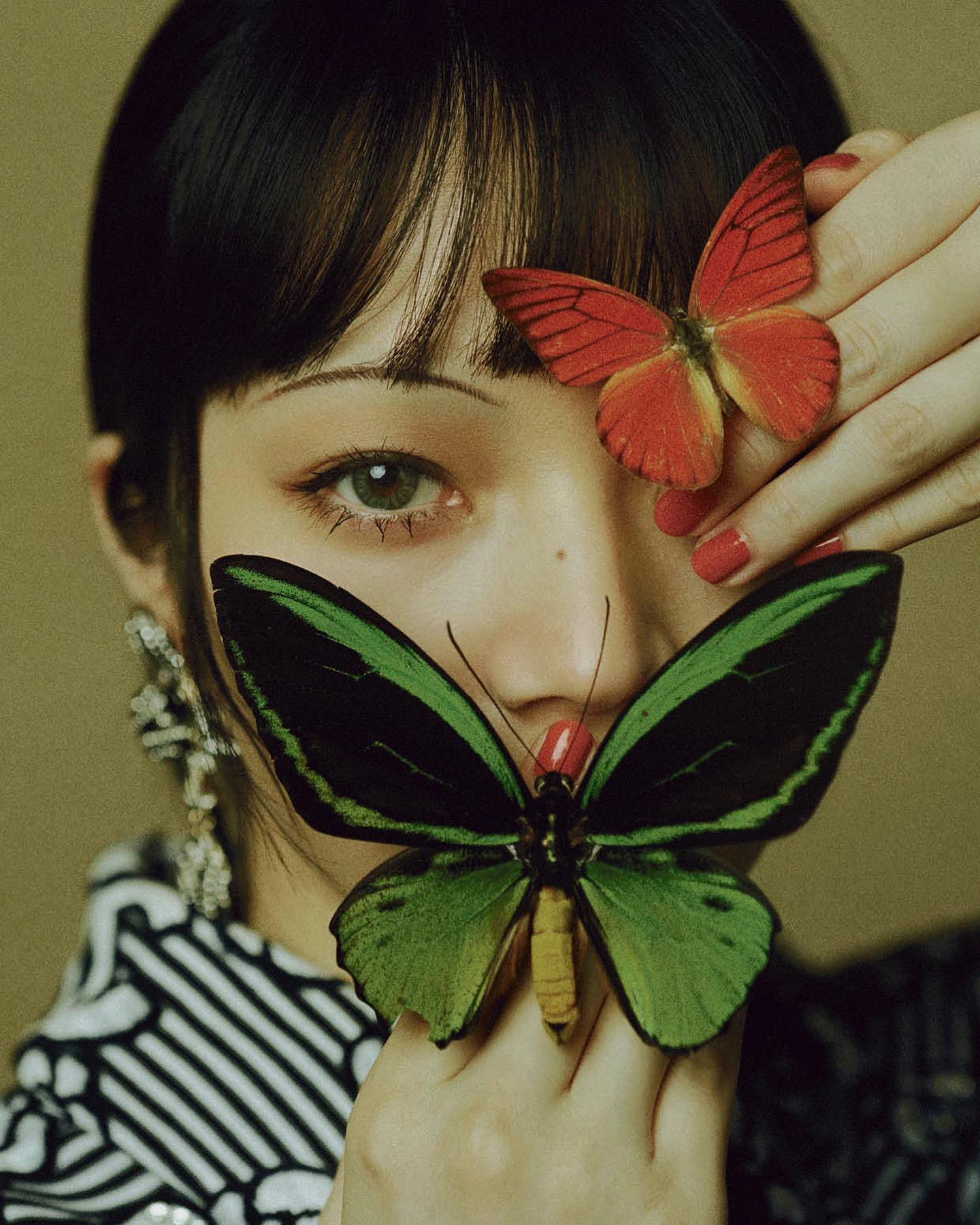 a closeup portrait of a woman with short, dark hair and bold makeup, holding two butterflies in front of her face. the top butterfly is red and black, partially obscuring her right eye. the bottom butterfly is predominantly green with black wings.  she wears a black and white patterned top. the background is a warm, neutral tone.