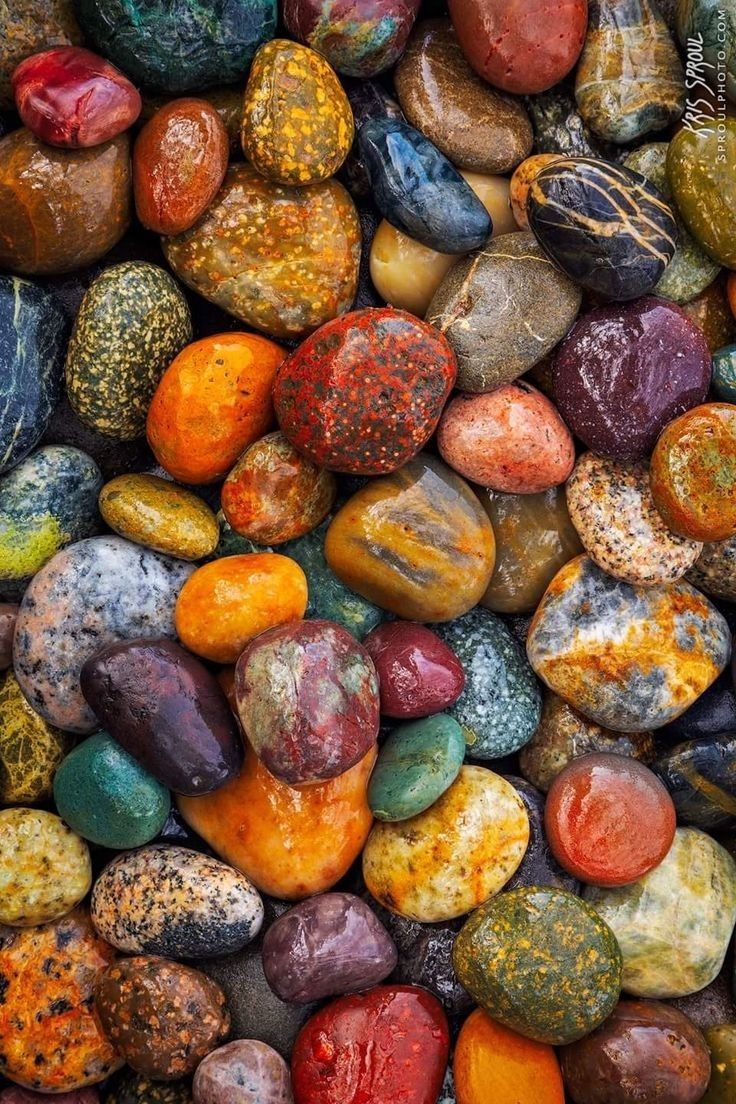 this photo shows a closeup view of colorful, smooth rocks and stones. the colors vary from reds and oranges to greens and blues, with many featuring unique patterns and markings. the rocks appear to be wet, reflecting light and creating a beautiful, abstract texture. the image is composed primarily of rocks, with only a hint of background visible. the photo evokes a sense of natural beauty and the wonders of the natural world.