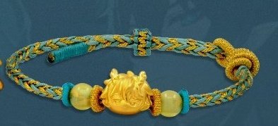 a closeup image of a gold and blue bracelet. the bracelet is made of braided blue and gold cord and has a gold charm in the middle. the charm is a stylized bull's head with horns and a nose ring. the bracelet is accented with three round gold beads and two round blue beads.