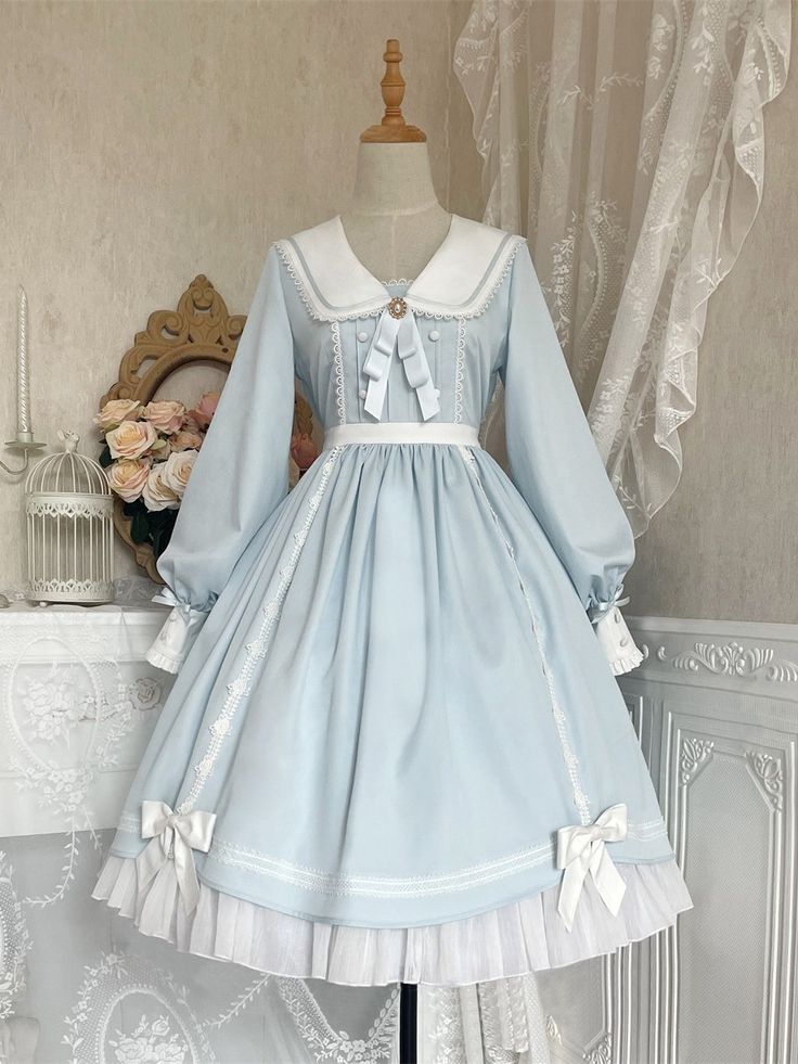 a baby blue lolita fashion dress is shown on a mannequin. the dress has a sailor collar, long sleeves, and a flowing skirt with lace trim. it is adorned with white bows and has a vintage, elegant feel.