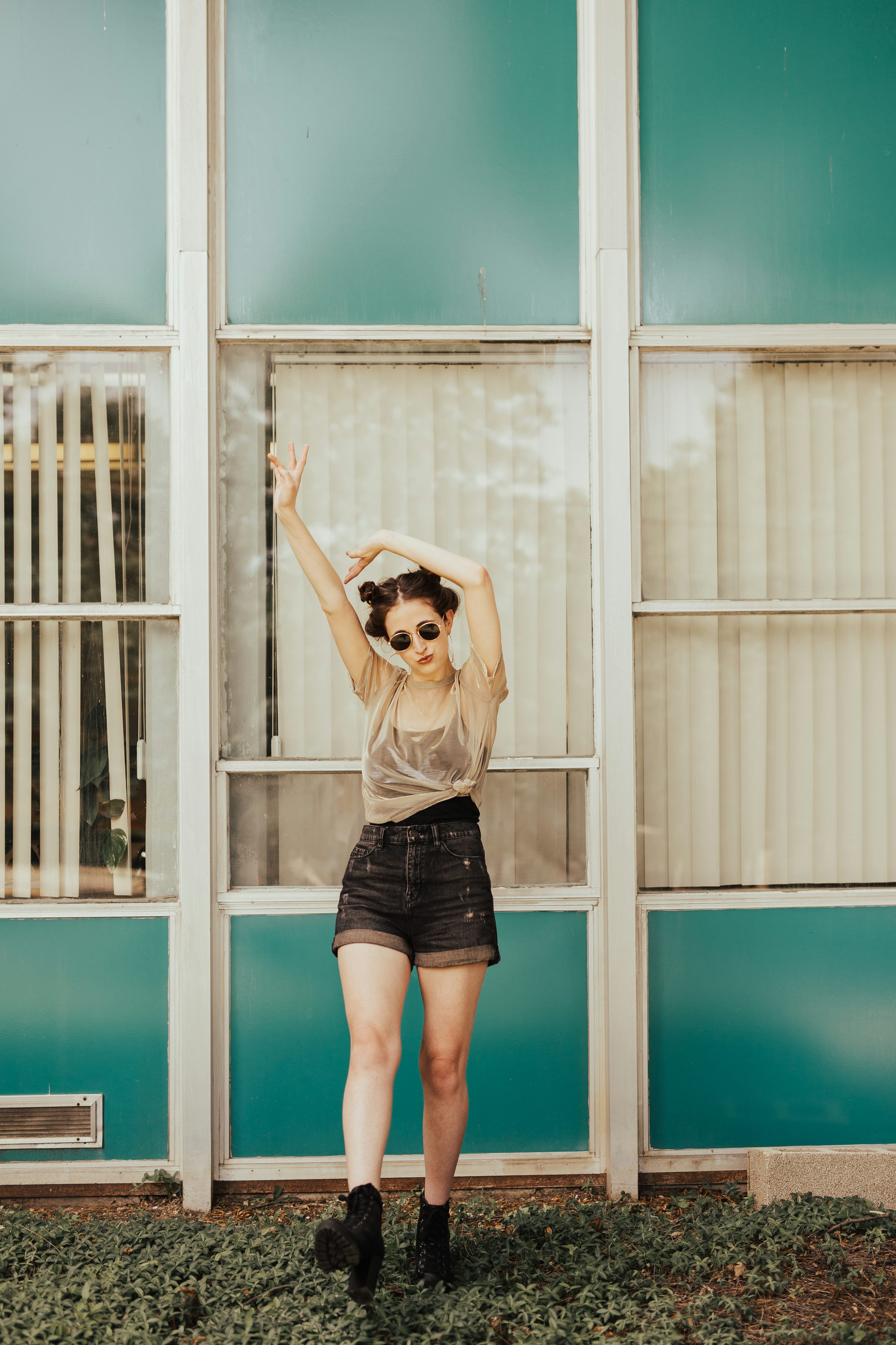 a fullbody shot of a young woman posing playfully in front of a teal and white building. she's wearing a sheer beige top, dark denim shorts, and black boots. her hair is styled in two buns, and she's wearing sunglasses. the background features large windows and a midcentury modern architectural style. the overall vibe is retro and fashionable. the image is welllit and has a vibrant color palette.