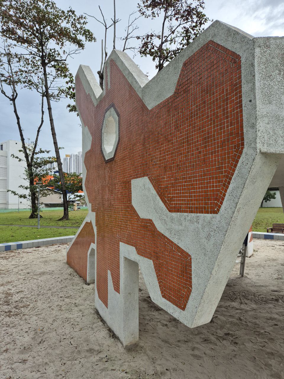 a concrete climbing frame with a red tiled section and a slide. the frame is in a sand pit and is surrounded by green grass. the frame is surrounded by trees in the background.