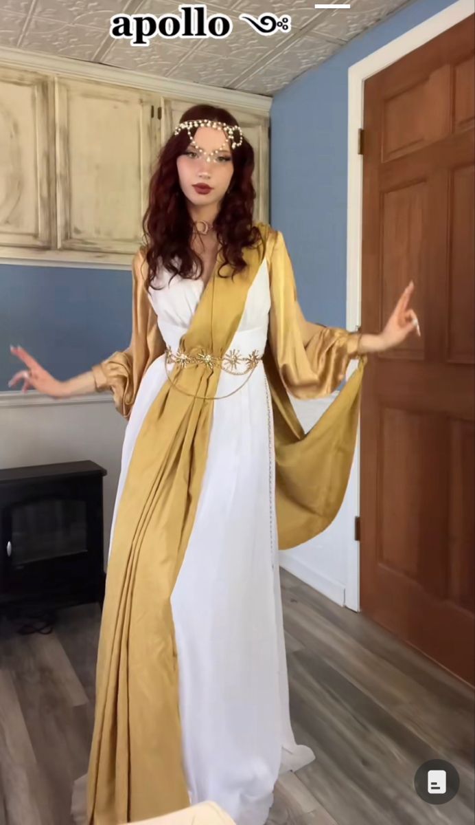 a woman dressed in a white and gold apollo costume. she is wearing a jeweled headdress and a gold belt. her hair is red. she is standing in a room with a door and a fireplace.