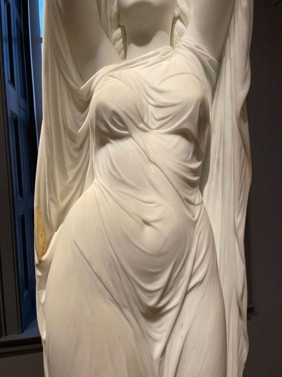 a closeup image of a white marble statue of a woman. the statue is draped in fabric that clings to her body, highlighting its curves. the statue is likely in a museum setting, as indicated by the lighting and background.