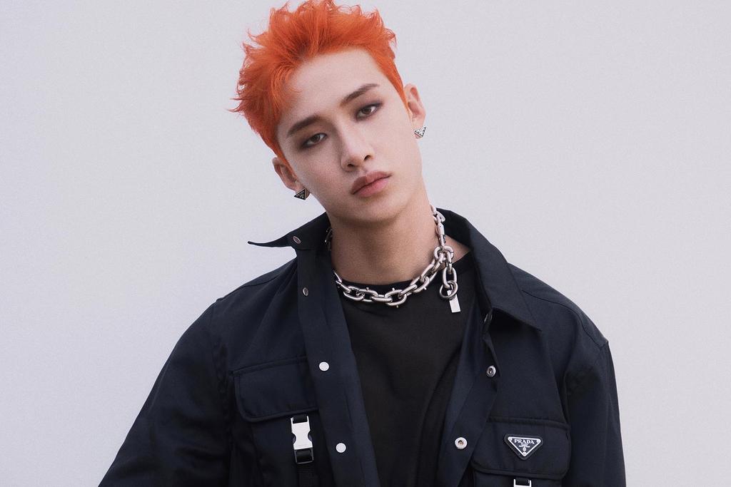 a young korean man with orange hair is wearing a black jacket, a silver chain necklace and looking directly at the camera. he is posed against a white background.