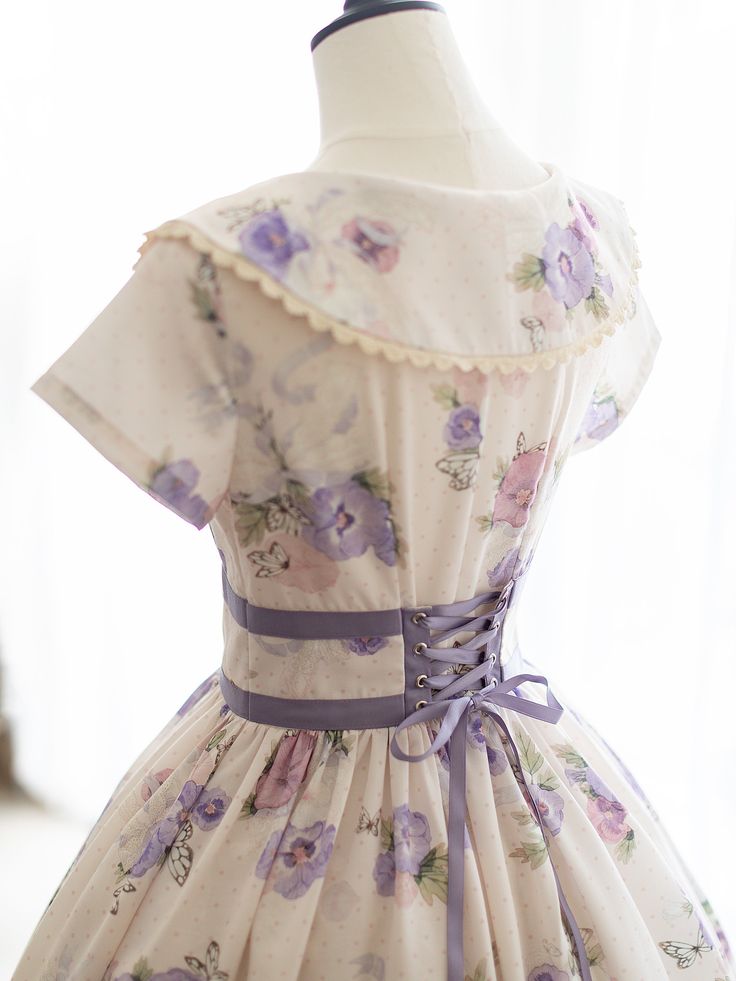 a back view of a dress on a mannequin. the dress is a light, flowy fabric with a floral print. it has a corset back with purple ribbon laces. the dress also features a large collar.