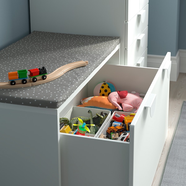 the image shows a white storage unit with open drawers filled with various children's toys. the toys include a toy train, stuffed animals, dinosaurs, and toy cars. the storage unit has a gray cushion on top. the setup is likely in a children's room or playroom, and the purpose is organization.
