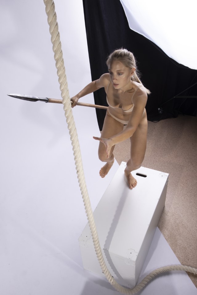 a woman with blonde hair stands barefoot on a white platform, holding a spear and reaching out as if grabbing a rope. the scene is set in a studio against a white background.