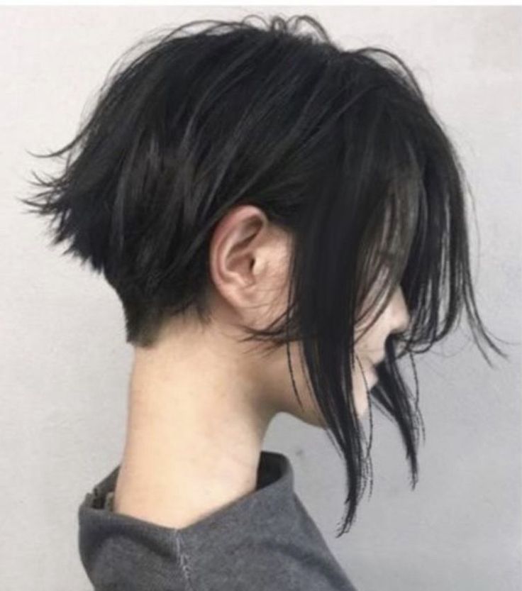 the image showcases a woman with a stylish short, black bob haircut. the bob is layered and textured, creating a slightly asymmetrical look with longer pieces framing the face.  a subtle nape undercut is visible at the back of her neck. her hair is styled with a side part, and some strands fall softly over her face. the overall style is modern and chic.  the woman is viewed in profile, looking down and to the side.  she is wearing a simple gray shirt. the background is a neutral light gray wall.