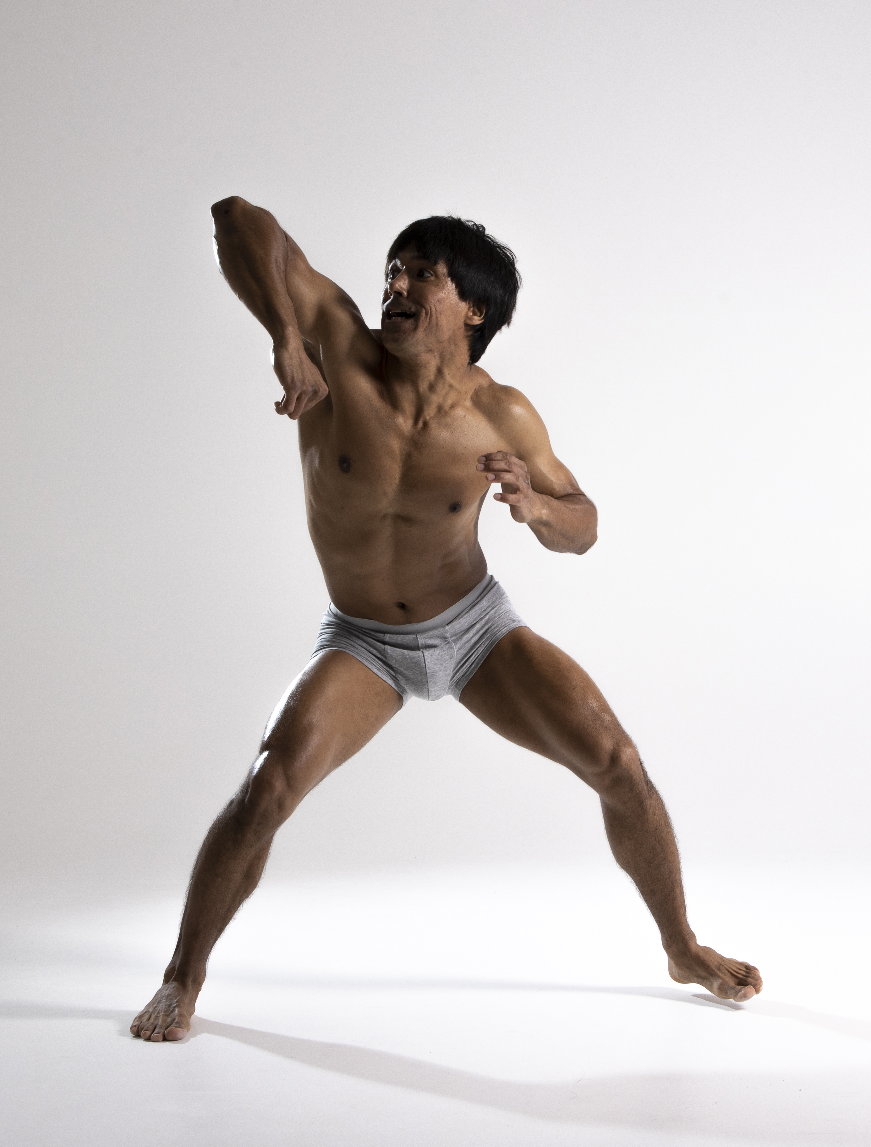 a muscular, shirtless man in gray underwear is posing barefoot in a studio setting. he is in an action pose, with one arm raised and his legs spread apart. the man has a toned and athletic build, suggesting physical fitness and a healthy lifestyle.