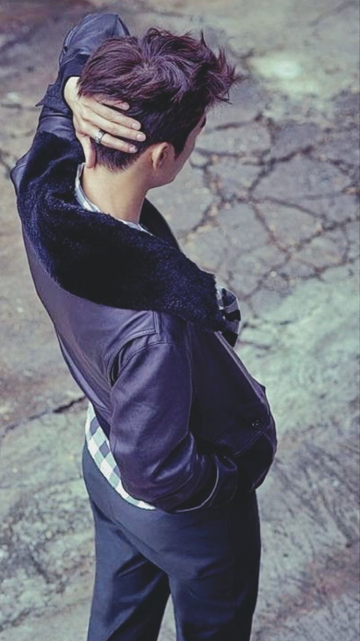 a young man in a dark leather jacket and pants is standing on a paved path with his hand on his face. he is looking away from the camera. the background is out of focus, but it appears to be a street scene with concrete ground.